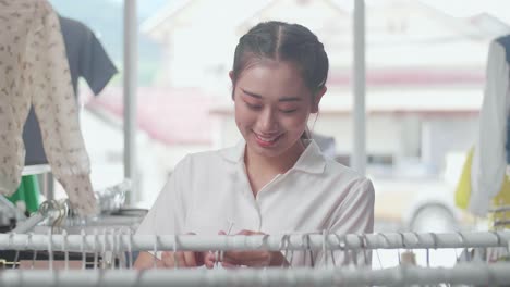 옷 가게의 아시아 여성 소유자가 가게의 재고를 확인하는 동안 레일에 드레스를 매달고 있습니다.