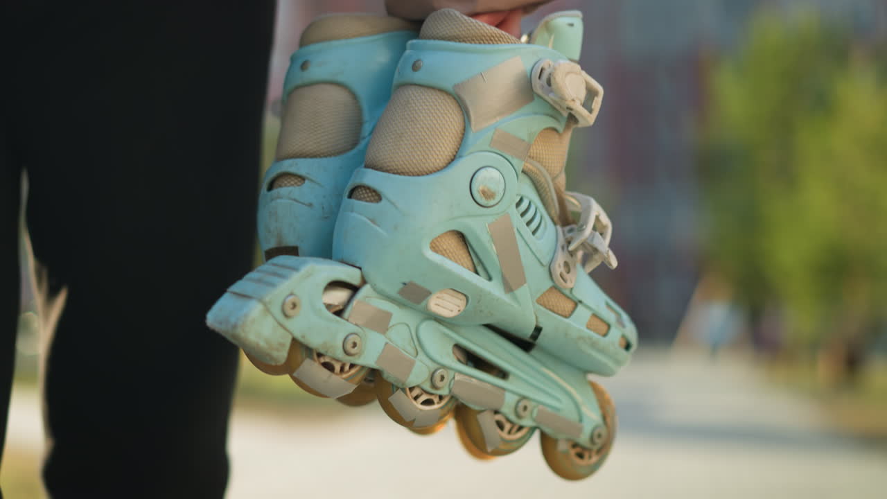 Close-up of a person holding a pair of rollerblades, wearing black trousers, walking through a park with a blurred background of trees and buildings