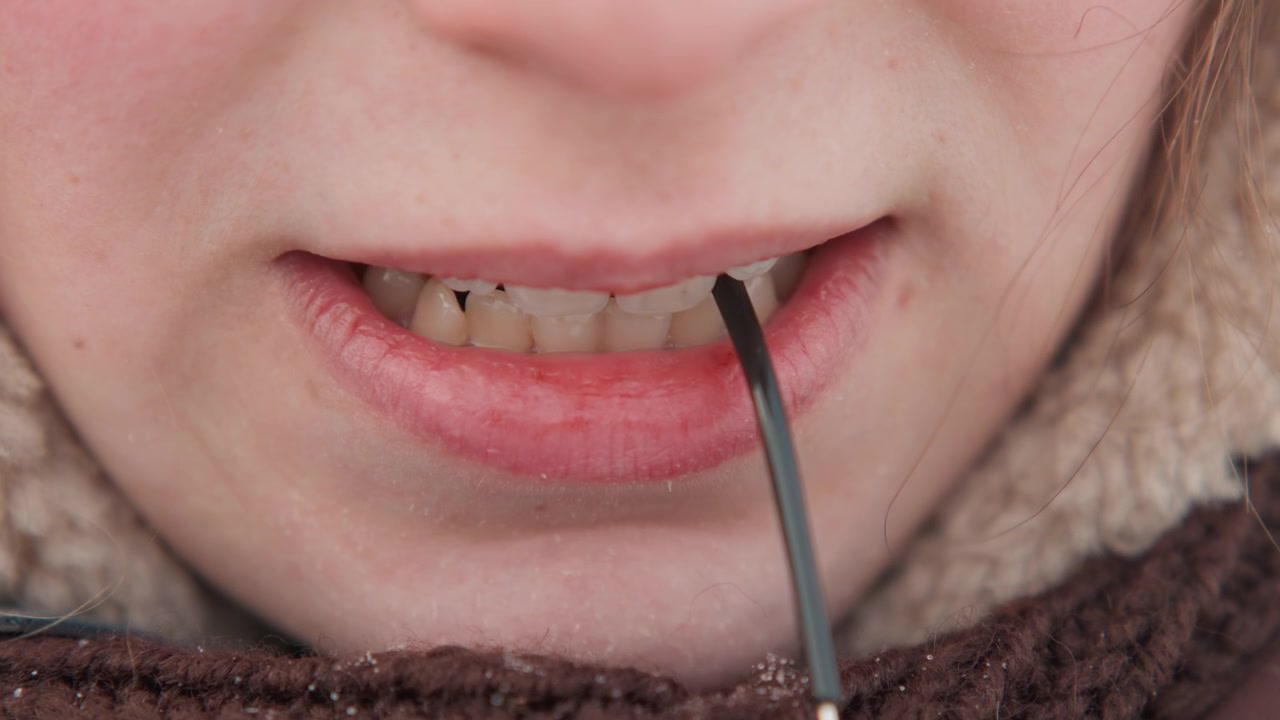 Vista frontale ravvicinata della parte inferiore del viso che tiene un sottile cavo nero tra i denti, tirando verso il basso con la bocca, poi rilassandosi in labbra chiuse, evidenziando la texture della pelle, i denti e una sottile espressione concentrata.