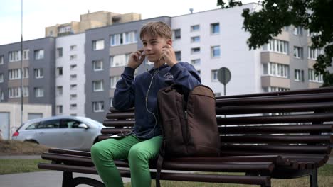 positive young man putting on earphones when sitting on the bench, mid shot