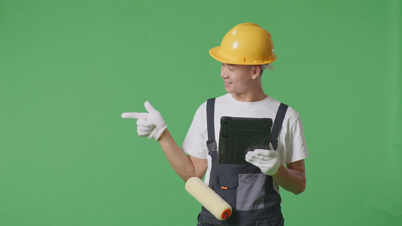Asian Man Painter Wearing Safety Helmet Using A Tablet And Pointing To Side While Standing In The Green Screen Background Studio