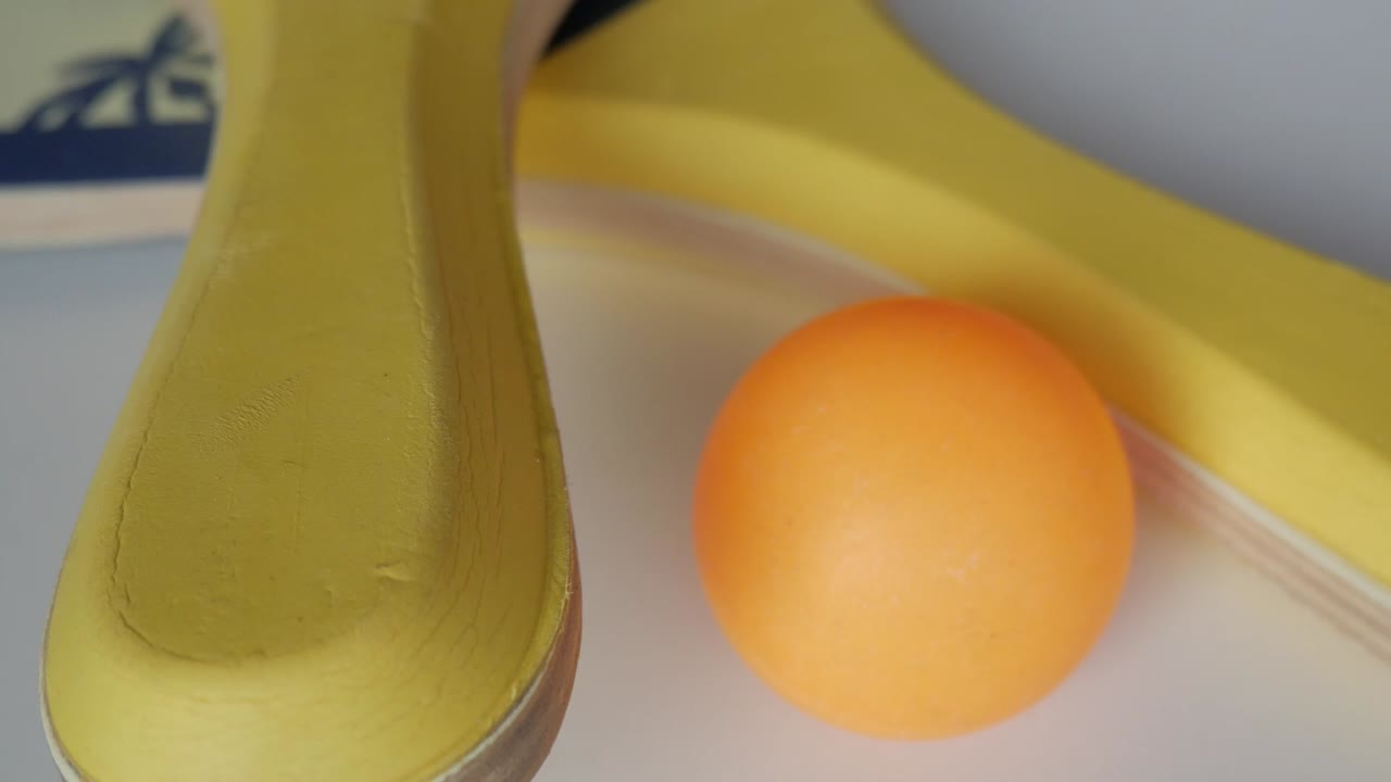 Close-up zoom out of a yellow beach tennis racket placed beside an orange ball on white background