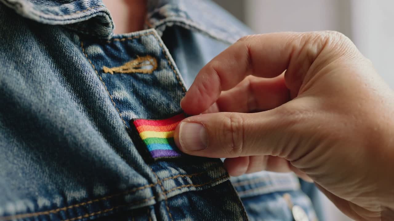 Entrando da destra, mano destra che pizzica e allinea una toppa arcobaleno sulla tasca di jeans per fissarla.