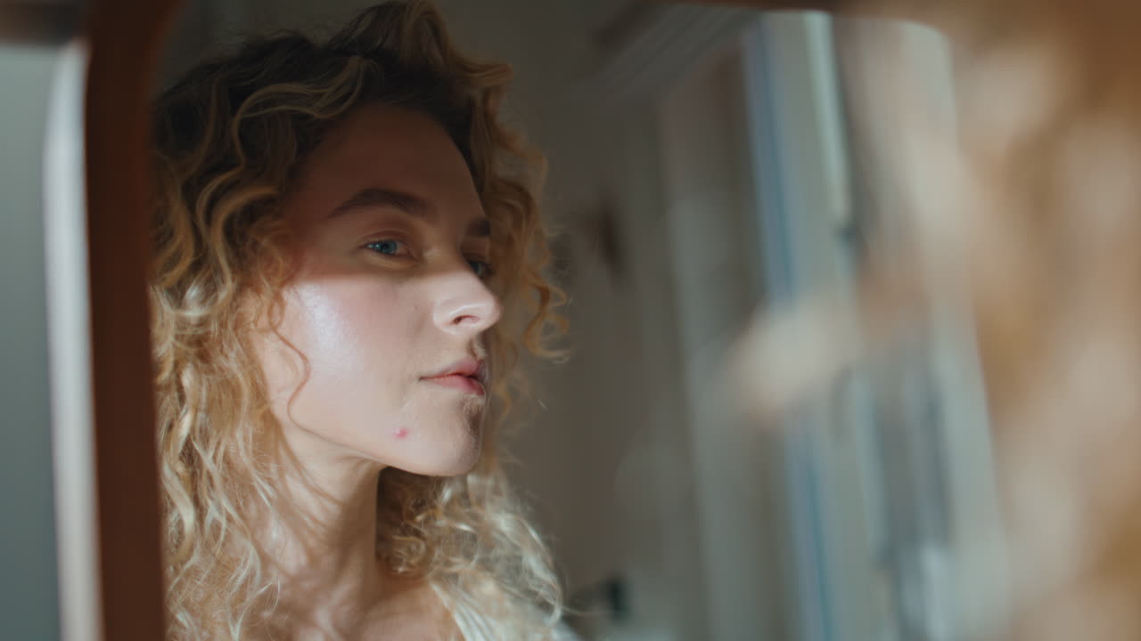 Primo piano di donna riccia che si guarda allo specchio nella luce del sole della stanza. Ragazza pensierosa che controlla la pelle.