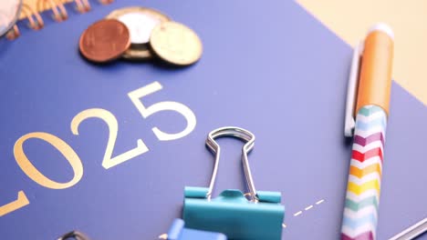 a blue planner with the year 2025 on it, along with a pen, coins, and other office supplies