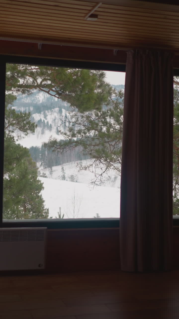 Big panoramic window of resort cottage house with view of scenic mountains and coniferous trees covered with snow. Winter landscape in Gorny Altai region