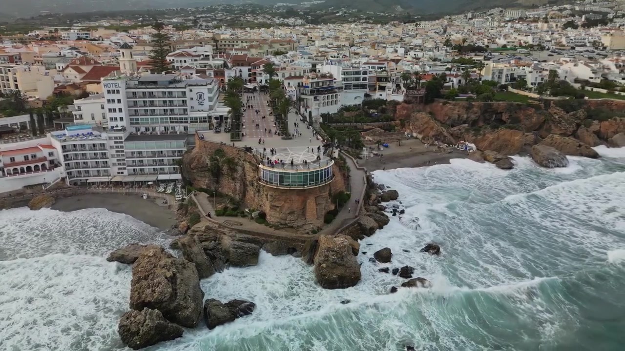 Luftdroneudsigt over Balcon de Europa udsigtspunkt på et klippefremspring med brusende bølger, palmeallé, hotelbygninger og en hvid andalusisk by, der strækker sig ind i landet, i overskyet dagslys.