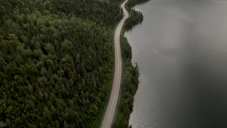 long and winding road by the lush forest and calm lake in gaspe peninsula, quebec, canada - aerial