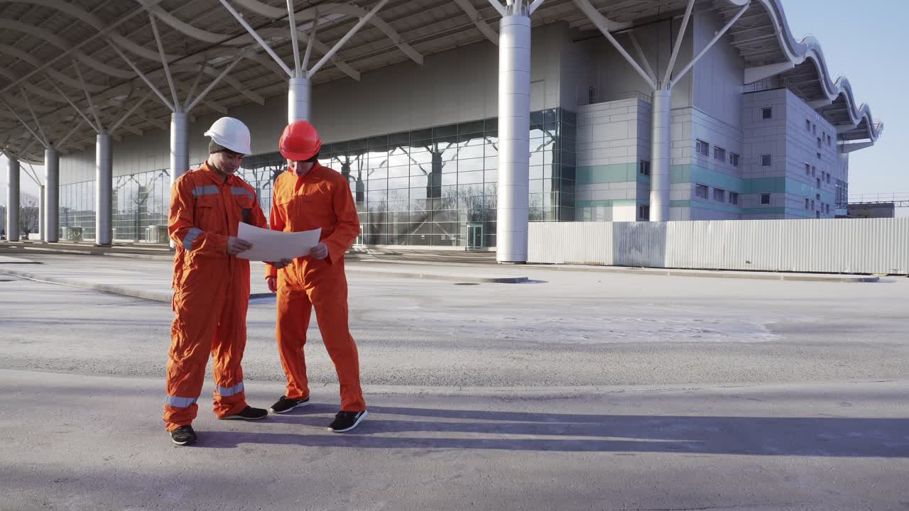 Two construction workers in orange uniform and helmets meeting each other at the bulding object, shaking hands and examining the constructed building together. Teamwork concept