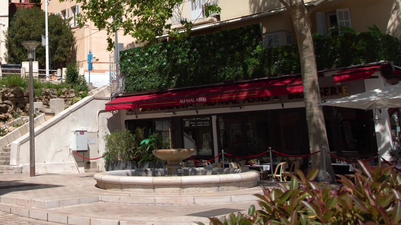Plano geral estático de um café mediterrâneo com toldos vermelhos e uma fonte de pedra