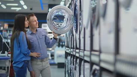 Young-married-couple-man-and-woman-in-appliances-store-choose-to-buy-a-washing-machine-for-the-house.-Open-the-door-looking-into-the-drum,-compare-the-design-and-characteristics-of-the-devices.