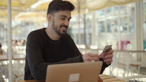 hombre sonriente con tarjeta de crédito y usando una computadora portátil en un café