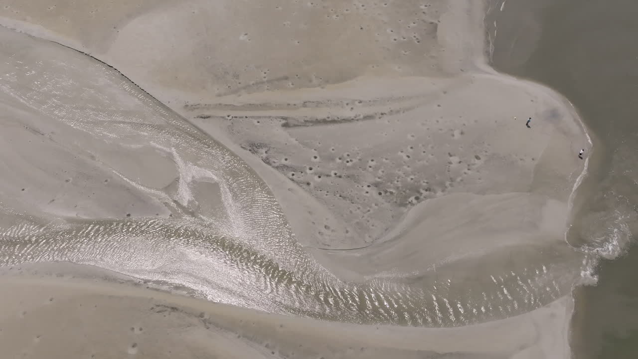 A winding water channel glistens in the sun, snaking through soft, pale sands with intricate ripples and tide-carved impressions