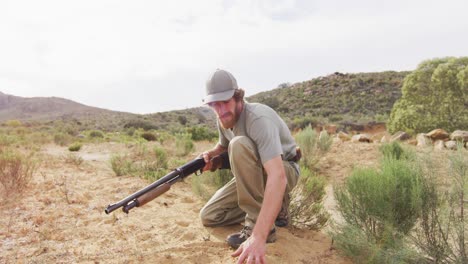 kneeling caucasian male survivalist holding hunting rifle, examining sandy terrain of wilderness