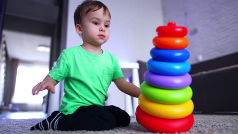 dark-haired toddler sitting on the floor in front of a toy pyramid. kid throws the pyramid taking away its basis.
