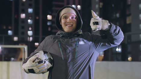 smiling man in hoodie alternating soccer ball in his hand while giving a thumbs-up with left hand raised above shoulder at night in urban setting