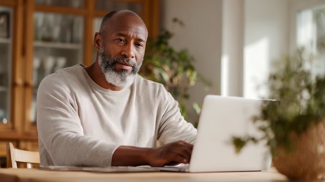 Focused Mature Man Engaged in Digital Work on Laptop at Home, Showcasing Concentration and Dedication to Tasks in a Comfortable Living Space with Warm Natural Light and Indoor Plants