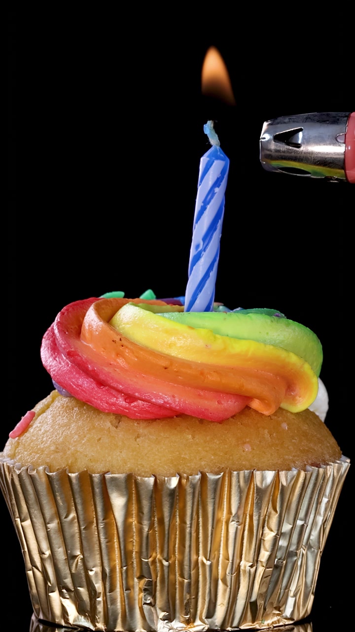 Un accendino accende una candela di compleanno a strisce blu su un cupcake glassato arcobaleno, il tutto su uno sfondo nero con illuminazione brillante e uniforme e ombre minime.