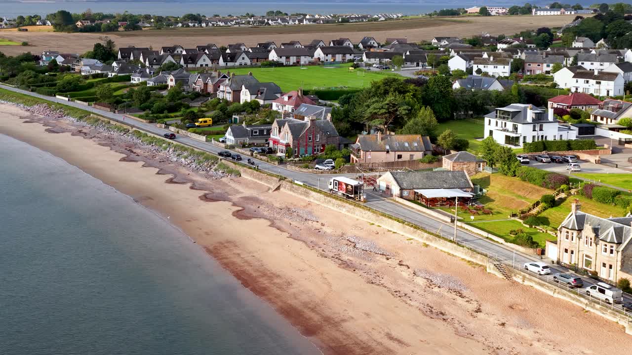 Drone glider over Rosemarkies kystlinje og afslører strandhuse, sandstrand og landligt landskab