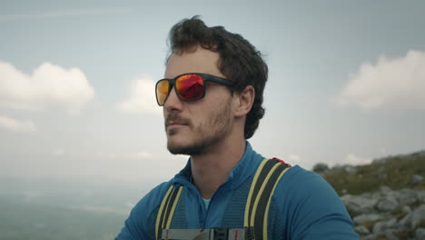 close shot of a hiker with a green backpack and dark sunglasses with red lenses looking at the beautiful view from peak of the mountain