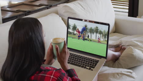 compuesto de una mujer sentada en casa sosteniendo un café viendo un partido de hockey en una computadora portátil