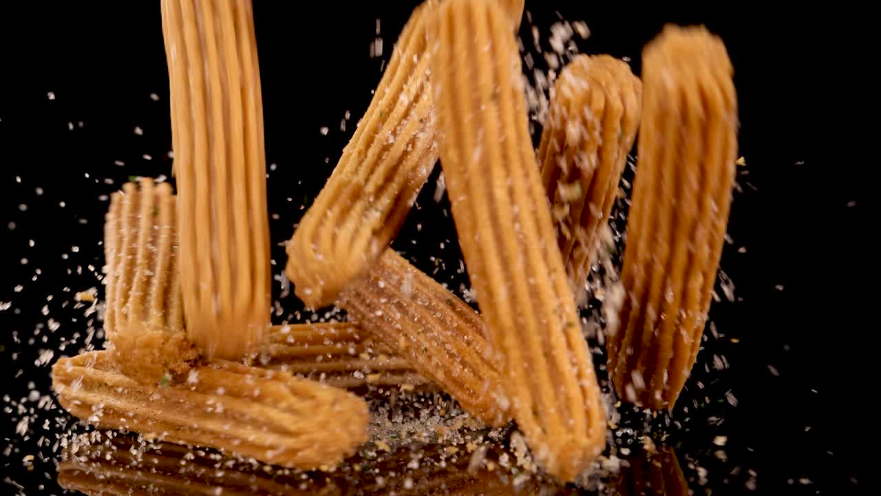 Golden churros fall and land on a sugar-coated surface, captured in slow motion with dramatic lighting and a clean black background for emphasis