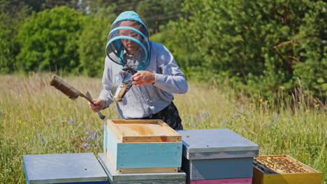 bee master on apiary. man in protective hat works with chimney in the beehive. beekeeper examining bees on a bee farm on green nature background.