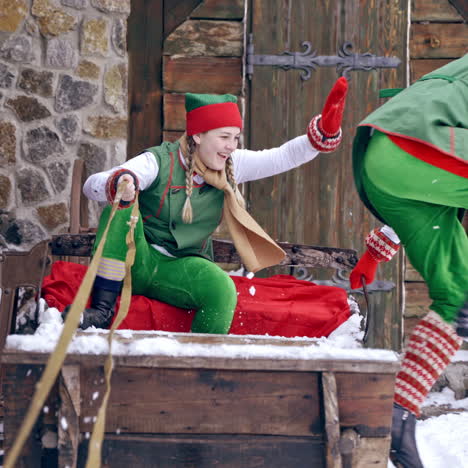 Two elves in green costumes sitting in wooden sleigh outdoors. Happy elves sit together in old fairy sleigh on the house background in winter. Christmas time.