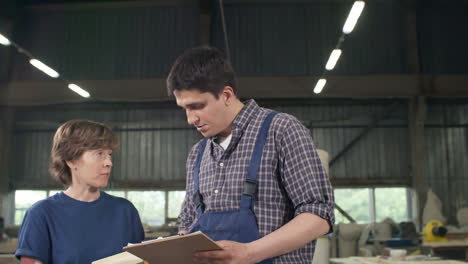 man and woman standing together in joinery and discussing wooden detail while their colleague carrying wooden plank on background