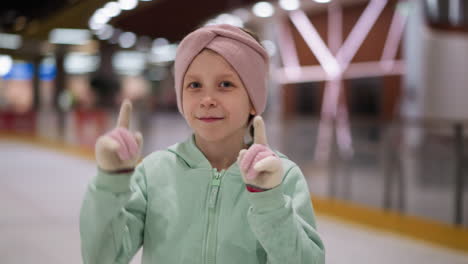 una vista cercana de un niño en un traje verde menta y una cinta rosa en la cabeza bailando lúdicamente en una pista de hielo, el fondo muestra a otras personas viendo