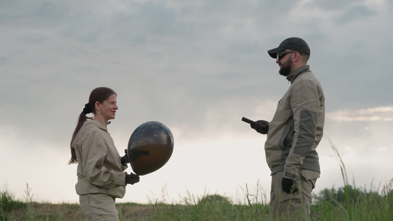 woman in protective jumpsuit holds black balloon while conversing with male colleague in open field wearing gloves and using device, dramatic cloudy sky backdrop creates moody