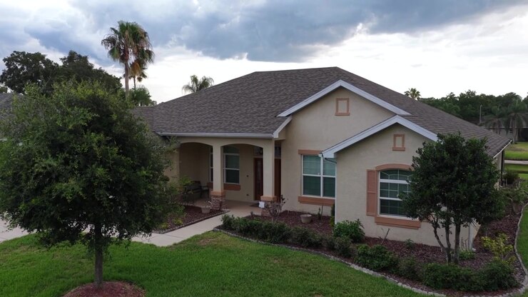 Casa a un piano in stucco con ingresso ad arco, situata dietro un prato curato, con un pick-up parcheggiato, vialetto, cassetta della posta, alte palme e scure nuvole temporalesche sopra. Plant City, Florida, Stati Uniti. Vista aerea.
