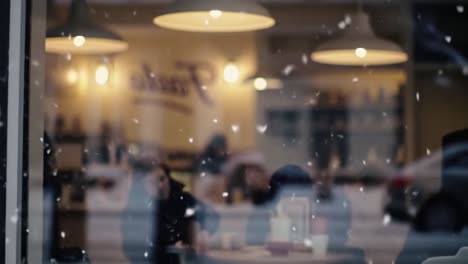 a cozy winter scene captured through a snowy window, featuring individuals inside a café enjoying their warm beverages while snow falls outside