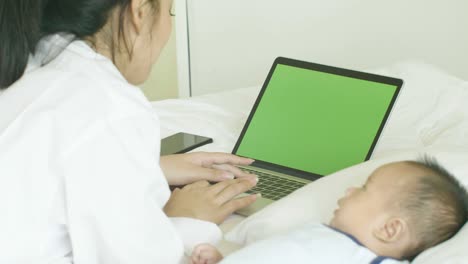 young asian mother with her newborn child working on greenscreen laptop in bedroom at home.