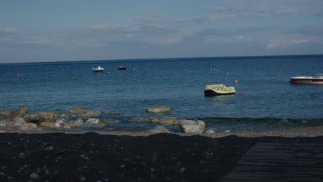 Boats-floating-in-sea-at-black-volcanic-sand-beach-in-Santorini,-Greece