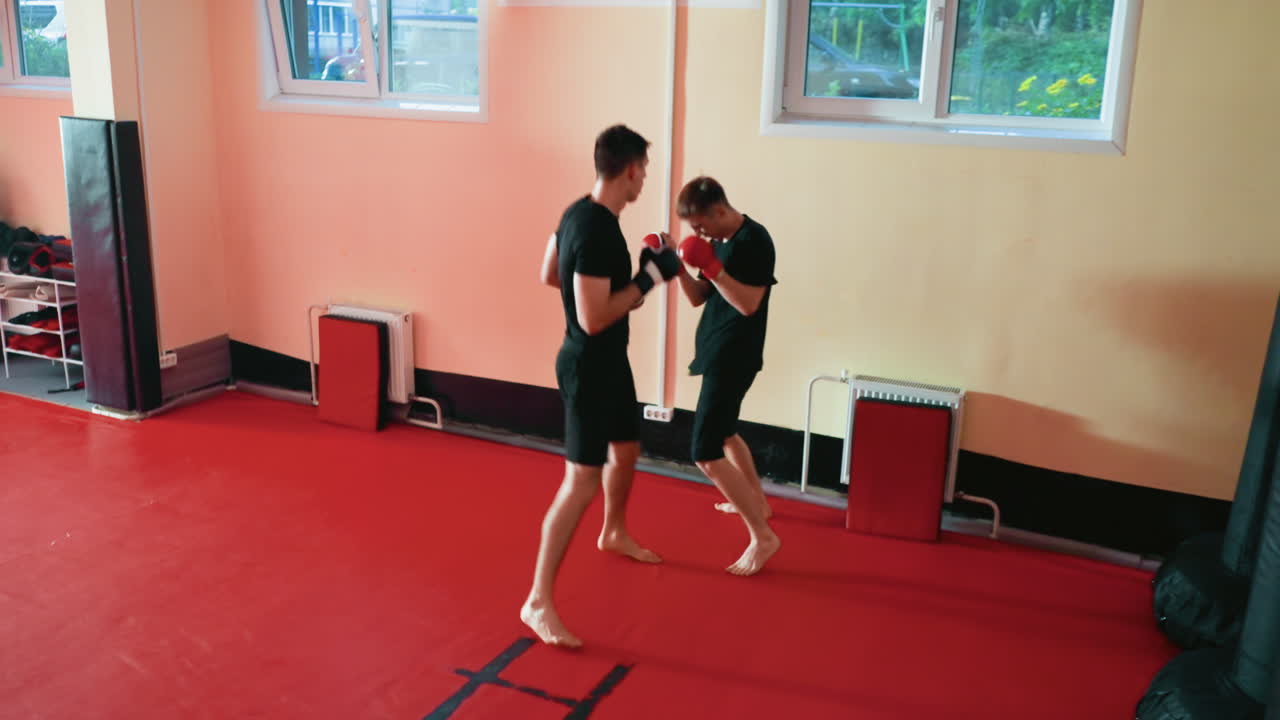 Boxers sparring barefoot in training gym, wearing gloves and black outfits, facing off with guarded stances on red mat, demonstrating agility, endurance, power, combat practice with determination