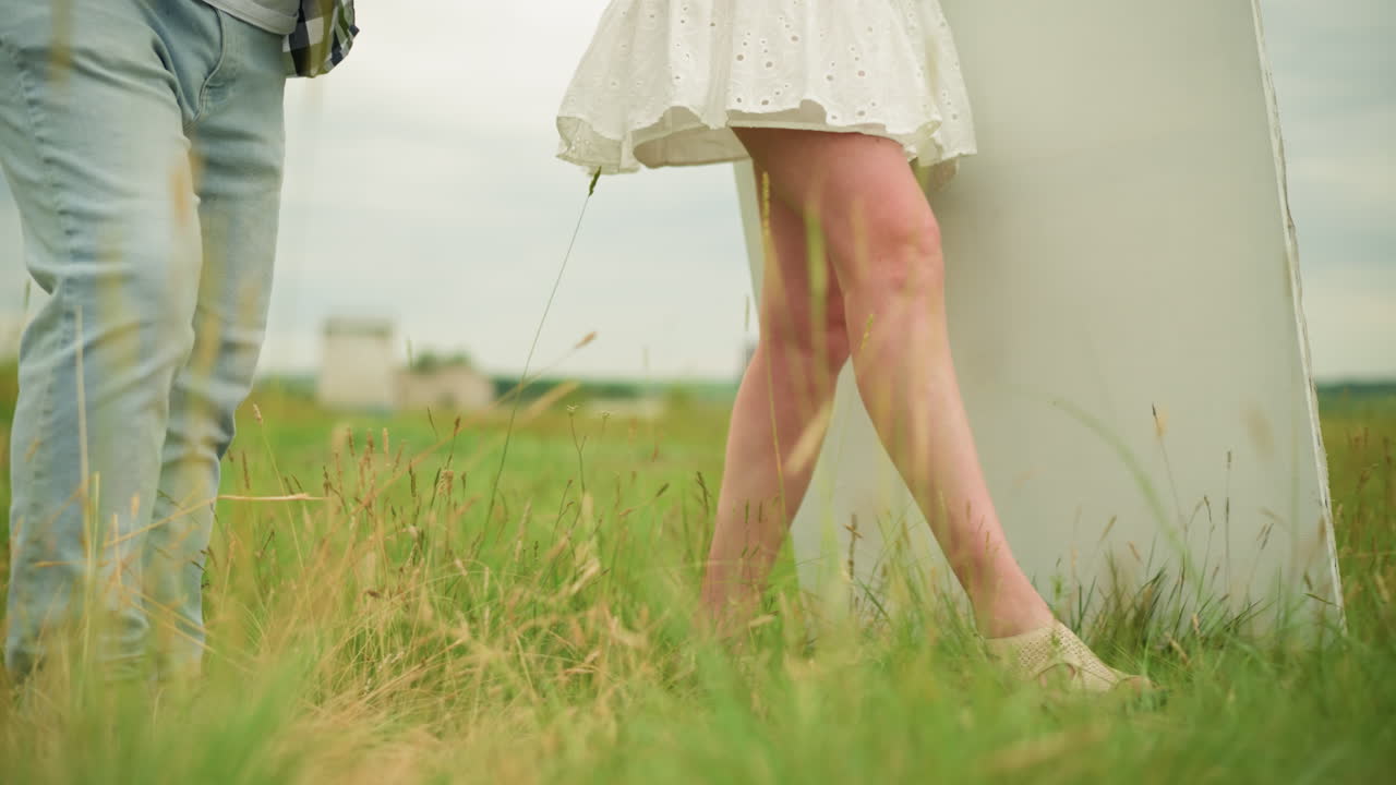 A front shot of a couple's legs walking through a grassy field. The man wears jeans and black shoes, while the woman, dressed in a short gown, walks beside him. natural, relaxed movement of the couple