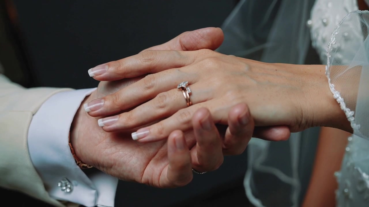 Holding bride's left hand, groom cupping and adjusting rings during ring exchange, beaded gown