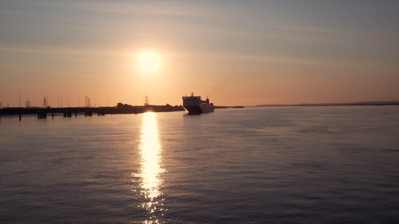 A cargo ship sails on the Thames River at sunrise, with the sun reflecting off the calm water. Industrial structures and power lines are visible in the background.