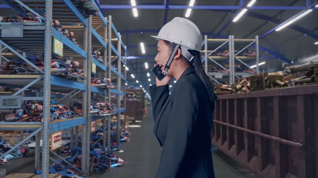 Vista laterale di una donna d'affari asiatica che parla al telefono mentre cammina in un magazzino industriale di rottami metallici e riciclaggio.
