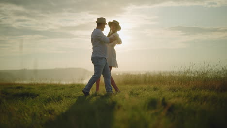 a loving couple dances on a sunlit grassy hill, enjoying a tender moment together. the man, dressed in a white shirt, hat, and jeans, embraces his wife, who is wearing a flowing white dress