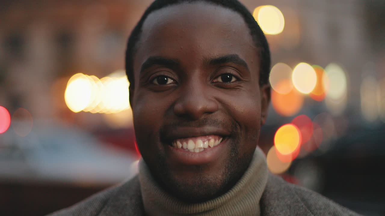 Close-up view of African American young man looking at the camera and smiling in the evening with city lights on the background