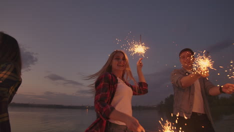 Cheerful-male-and-female-friends-are-running-along-the-beach-at-sunset-holding-sparkling-fireworks-and-runaway-lights-in-slow-motion.-Dancing-and-sunset-party-on-the-beach