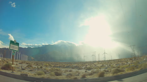 sun rays shine over the desert as wind turbines spin at the base of a mountainside. located in joshua tree california.