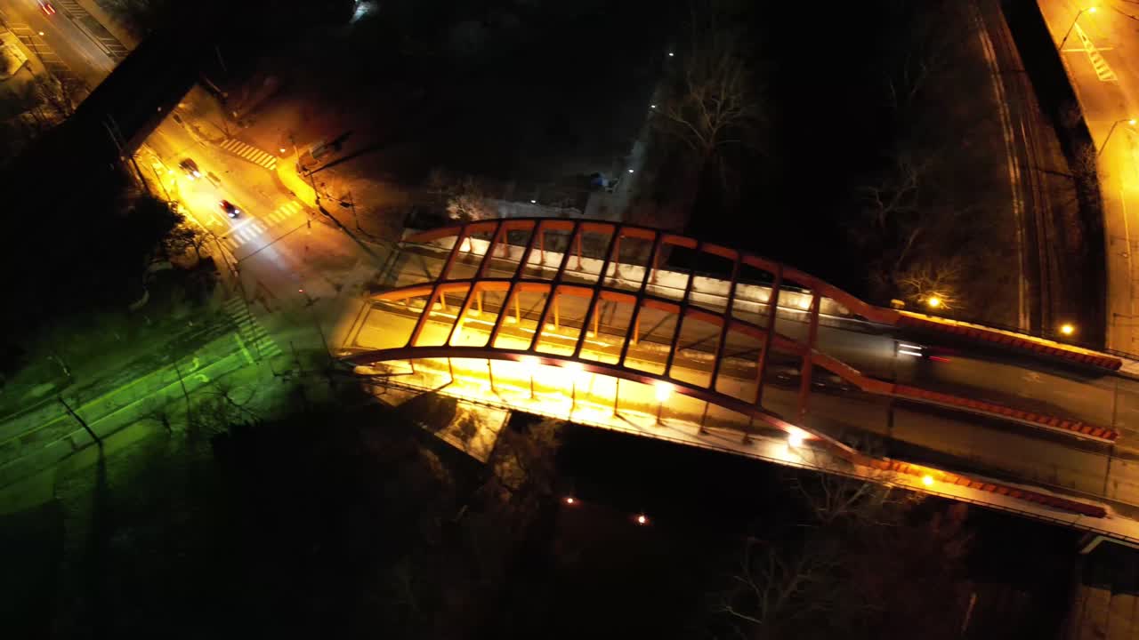 A rotating aerial timelapse over a glowing pedestrian bridge in Youngstown, Ohio at night, showcasing vivid lighting and motion blur from traffic below.