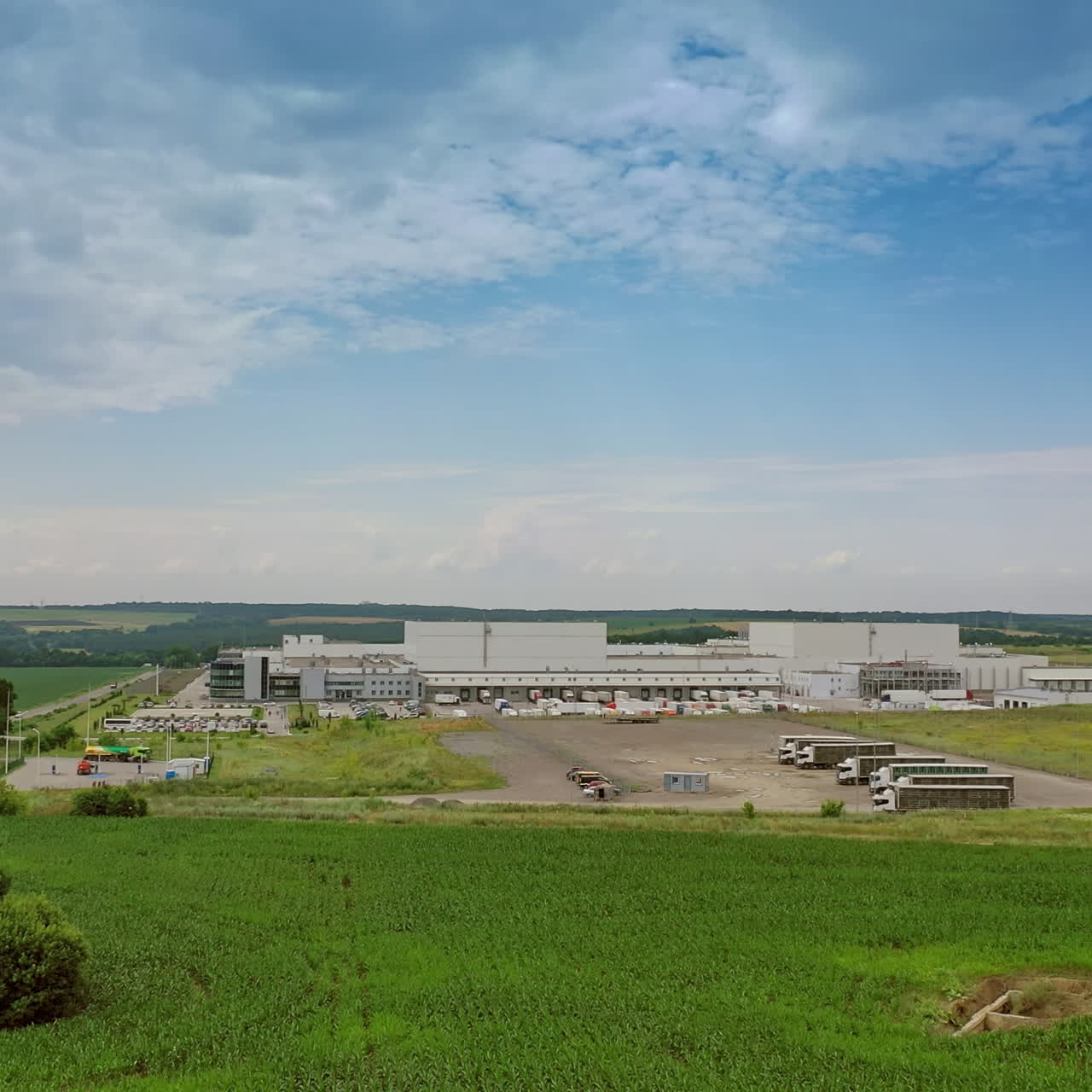 Exterior of industrial complex in the countryside. Modern buildings with parking for trucks among green fields under blue sky in summer.