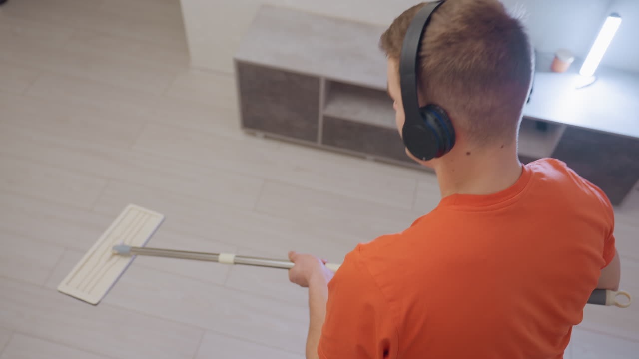 Rear angle view of man in orange shirt wearing headphones mopping wooden floor while enjoying music, focusing on cleaning task with modern home interior background