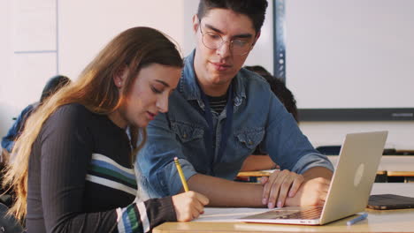 Male-Teacher-Giving-One-To-One-Support-To-Female-Student-Working-At-Desk-On-Laptop