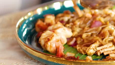a detailed view of shrimp pad thai being served with a spoon and fork on a decorative plate.