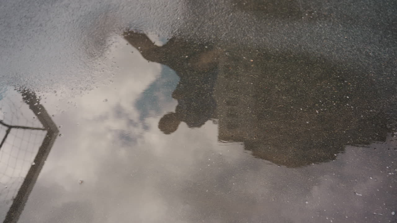 Pfützenreflexion eines Fußballers im Regen, nasser Asphalt und Regentropfen erzeugen filmische Stimmung, Torpfosten-Silhouette rahmt Spieler ein, Stiefel trifft Ball mit Spritzer, Wellen und Wolken spiegeln sich auf der Oberfläche
